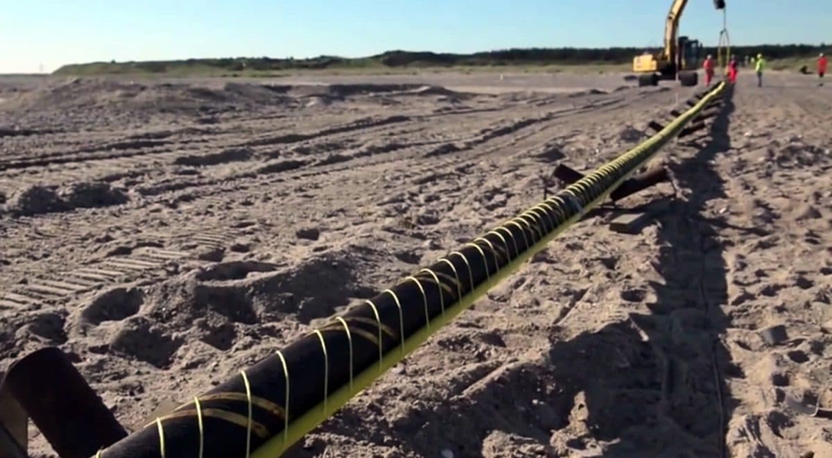 En lang kabel blir installert på en sandstrand, med anleggsutstyr og arbeidere i bakgrunnen.