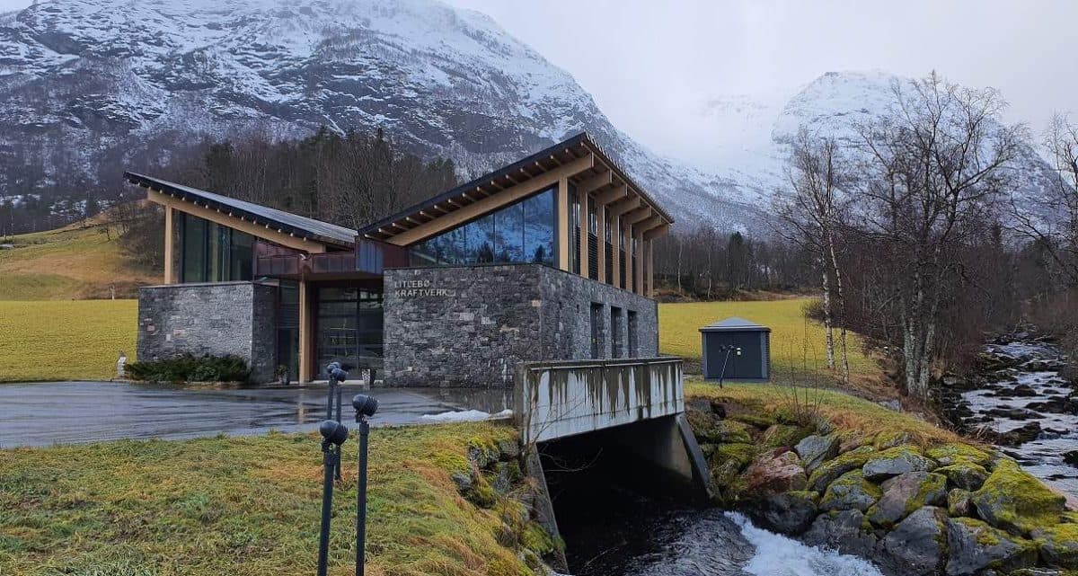 Moderne bygg med stein- og glassfasade, beliggende ved en bro over en liten bekk, med snødekte fjell og vinterlandskap i bakgrunnen.