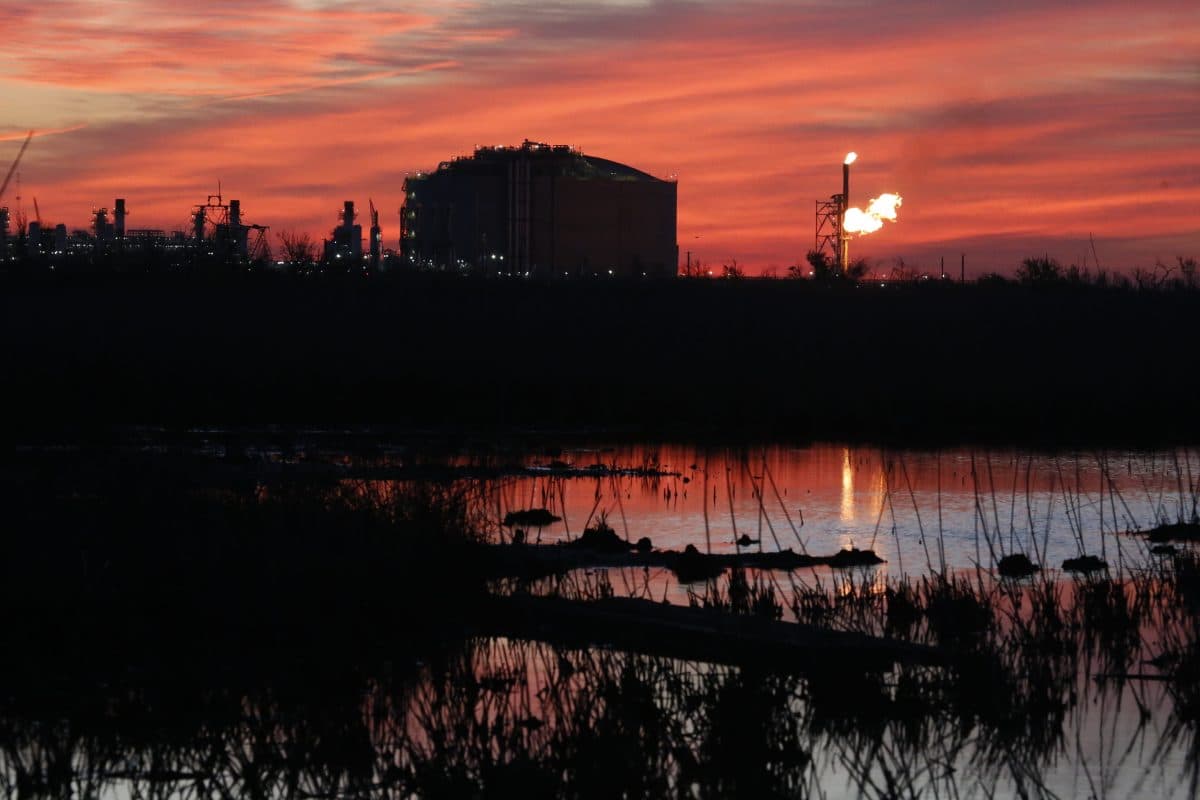 Industrianlegg med gassfakkel er silhuett mot en rød-oransje solnedgangshimmel. Scenen reflekteres i en vannmasse i forgrunnen.