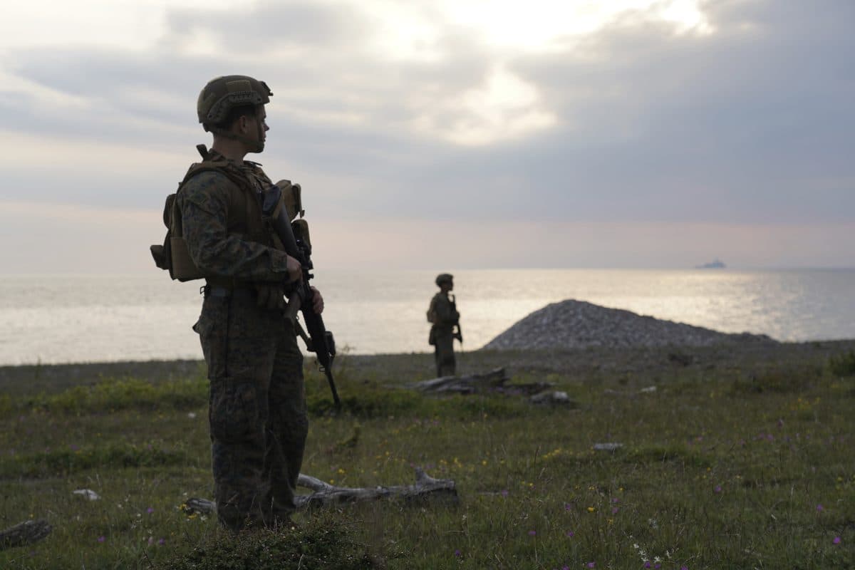 To soldater i militærutstyr står på en gressmark nær sjøen, med en soldat i forgrunnen og en i bakgrunnen under en overskyet himmel.