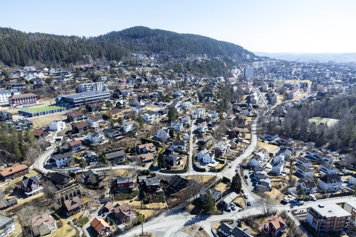 Luftfoto av et forstadsområde med en blanding av hus og trær, svingete veier og et kupert landskap i bakgrunnen.