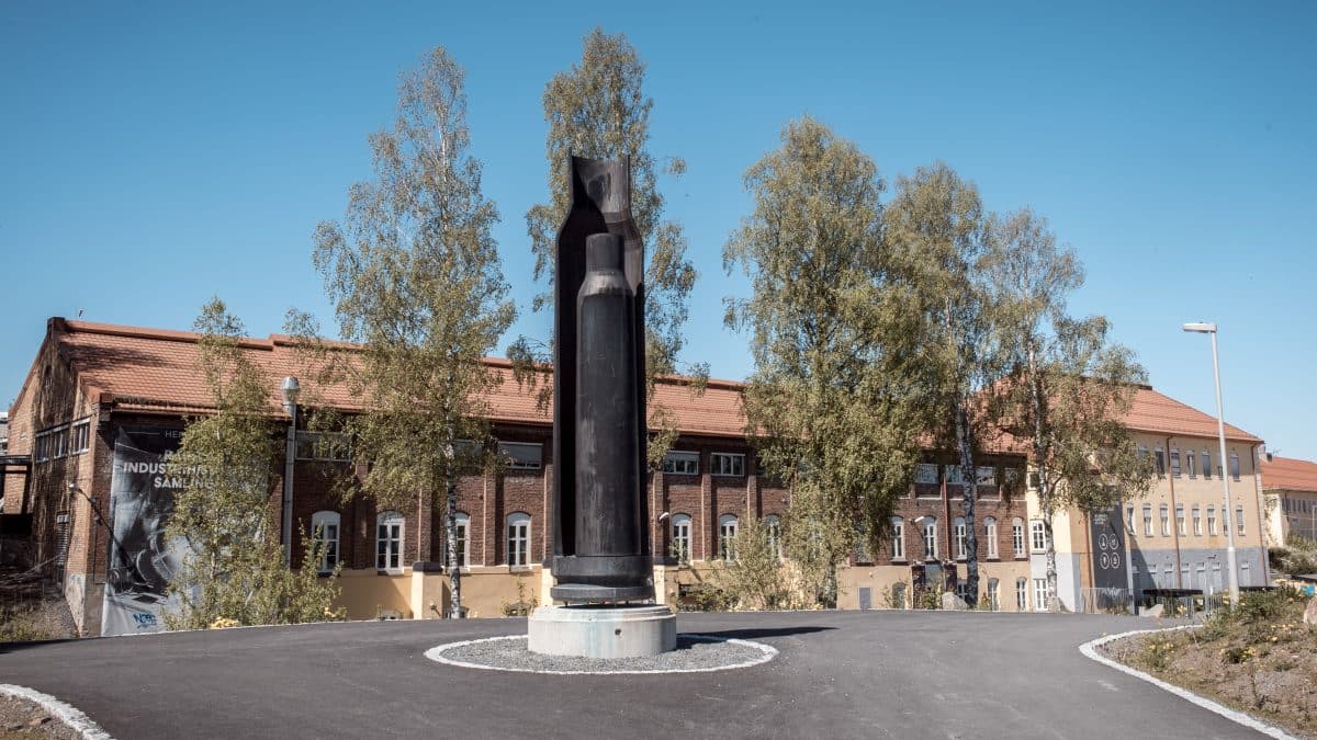 Høy, abstrakt metallskulptur foran en murbygning med trær og en klar blå himmel.