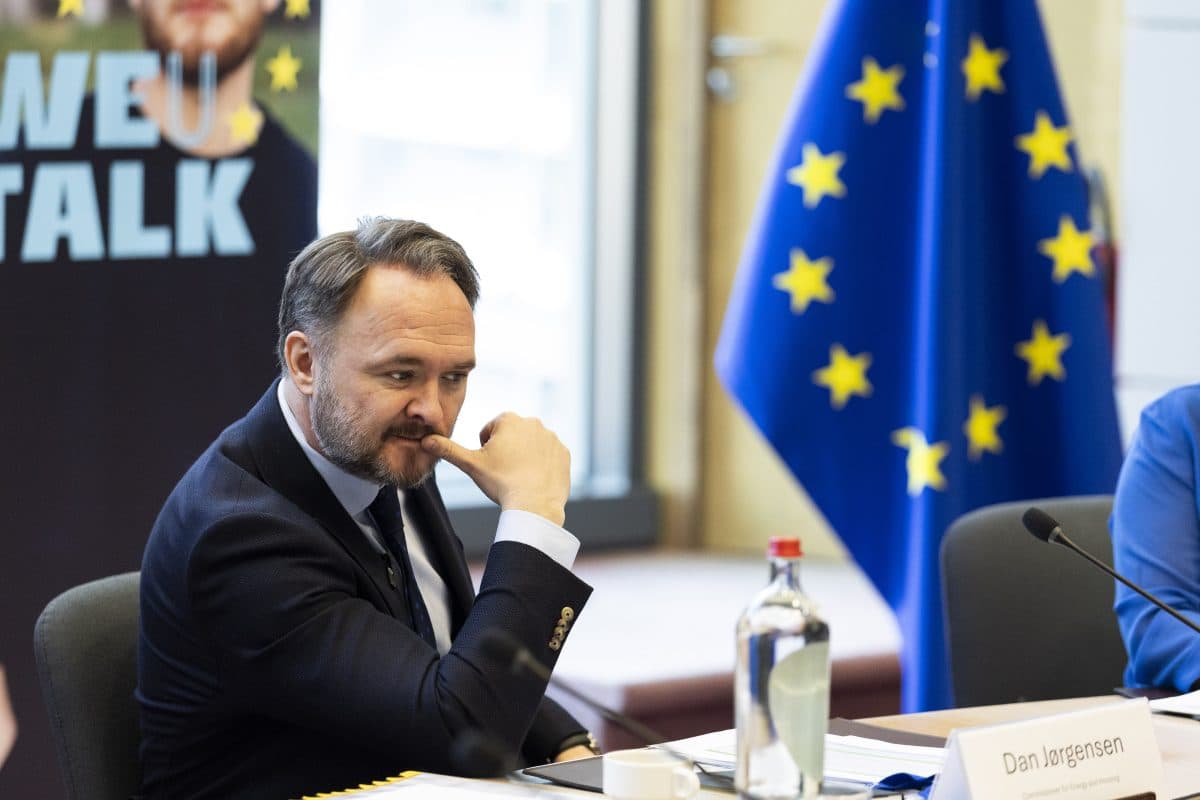 En mann i dress sitter ved et konferansebord med et EU-flagg i nærheten. Han legger hånden på haken og ser ettertenksom ut. Et skilt og en flaske vann står på bordet.