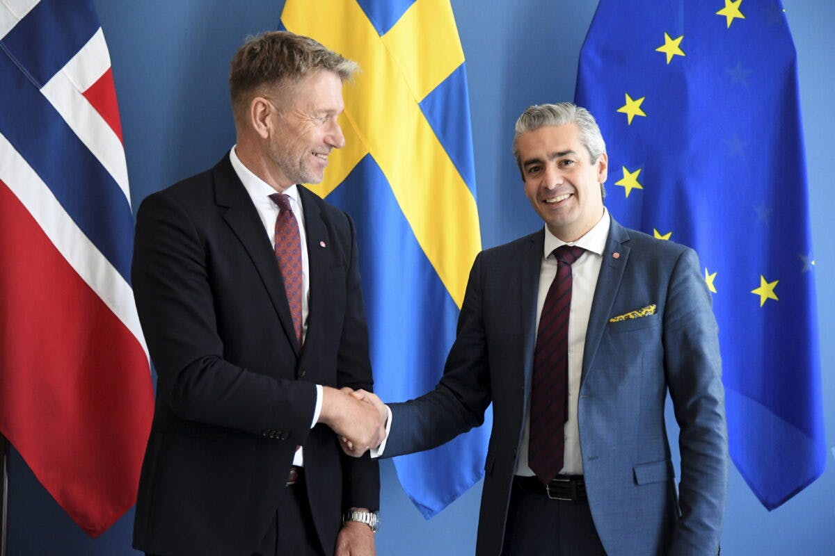 Terje Aasland tar Khashayar Farmanbar i hånden med norsk, svensk og EU-flagg i bakgrunnen.