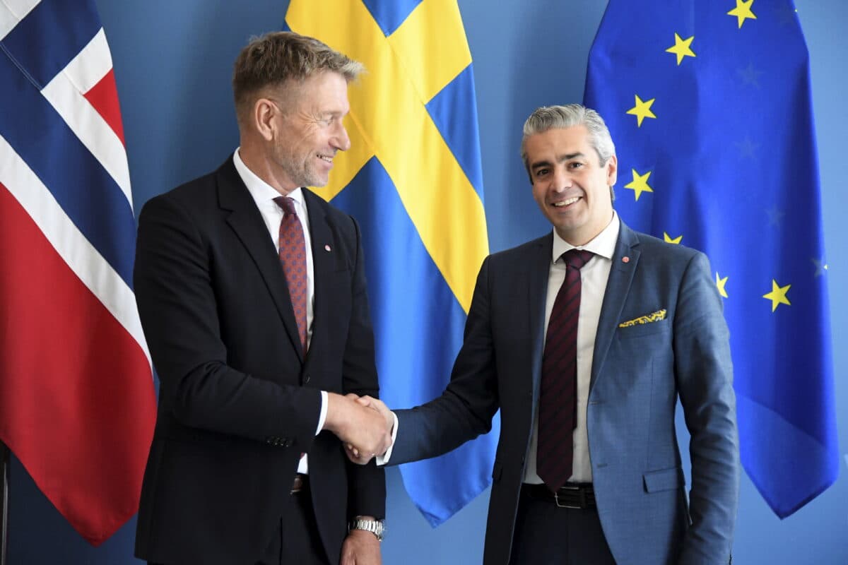 Terje Aasland tar Khashayar Farmanbar i hånden med norsk, svensk og EU-flagg i bakgrunnen.