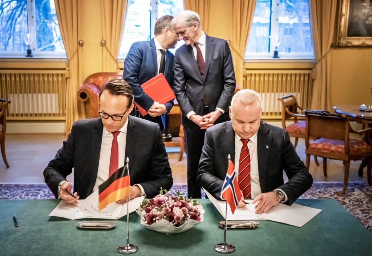 Opedal og Krebber signerer avtale ved et bord, Habech hvisker Støre noe i øret i bakgrunnen.