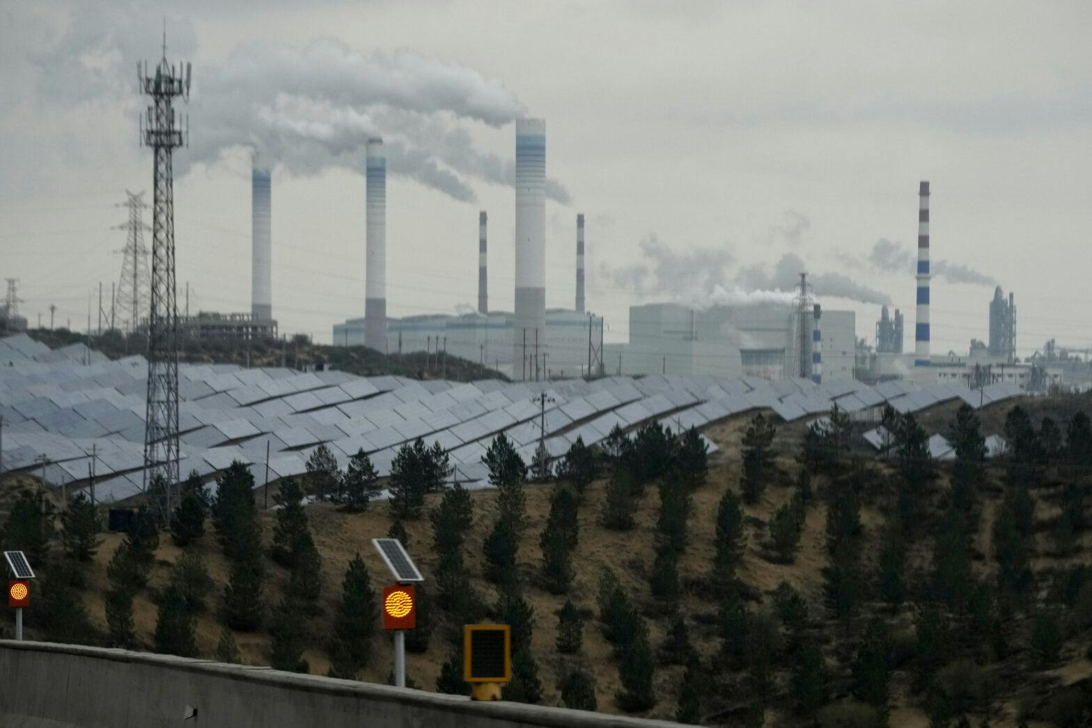Et bilde av en solcellepark foran et industriområde med rykende piper.