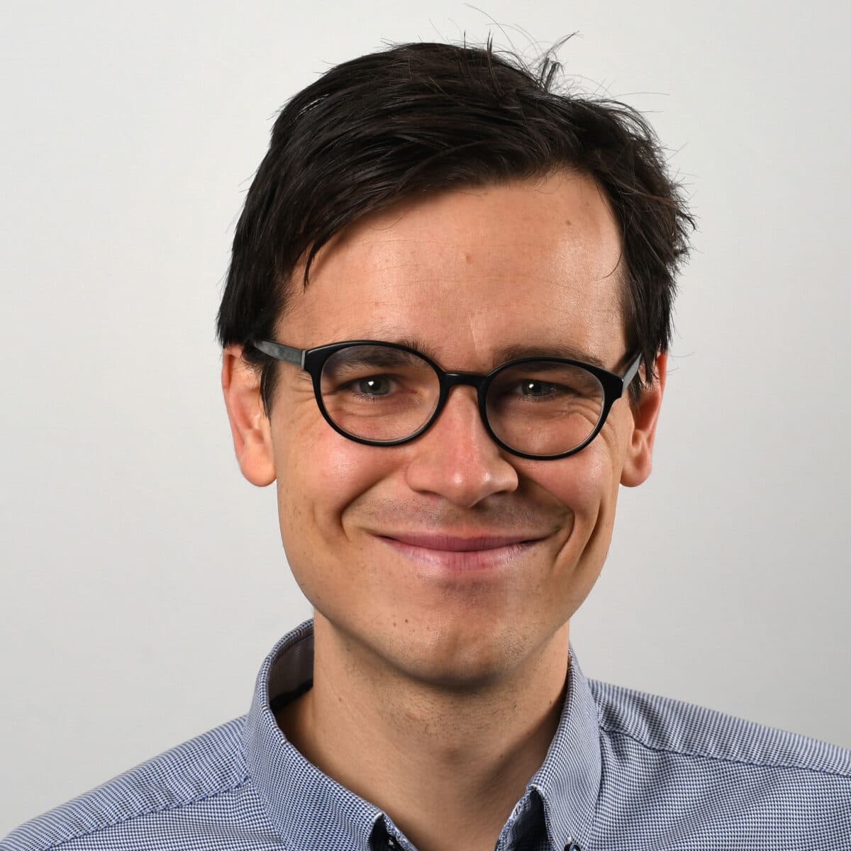 En mann med briller smilende foran en hvit bakgrunn.