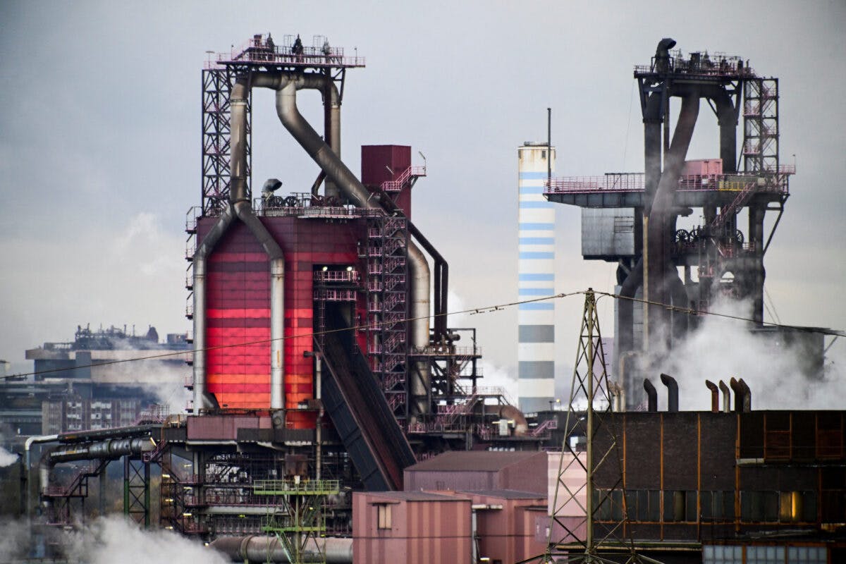 Industriell stålfabrikk med store røde ovner og røykstenger som avgir damp og røyk mot en overskyet himmel.