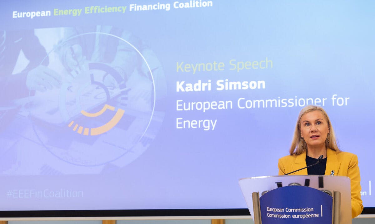 En kvinne står på et podium og holder en hovedtale på en konferanse med et banner om europeisk energieffektivitetsfinansieringskoalisjon i bakgrunnen.
