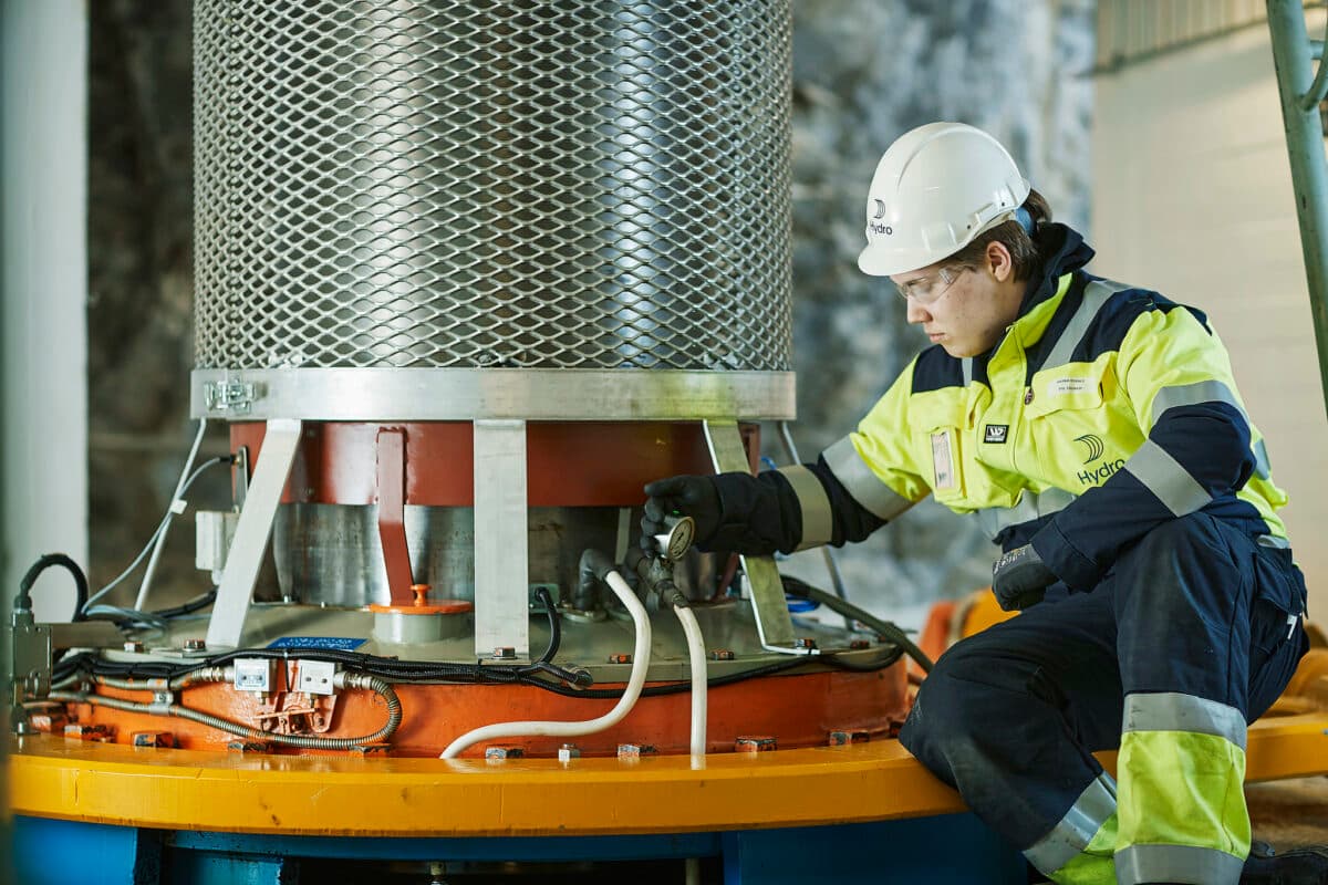 En arbeider i synlig utstyr og en hjelm inspiserer industrielt utstyr i et anlegg ved å bruke en lommelykt for å undersøke detaljer.