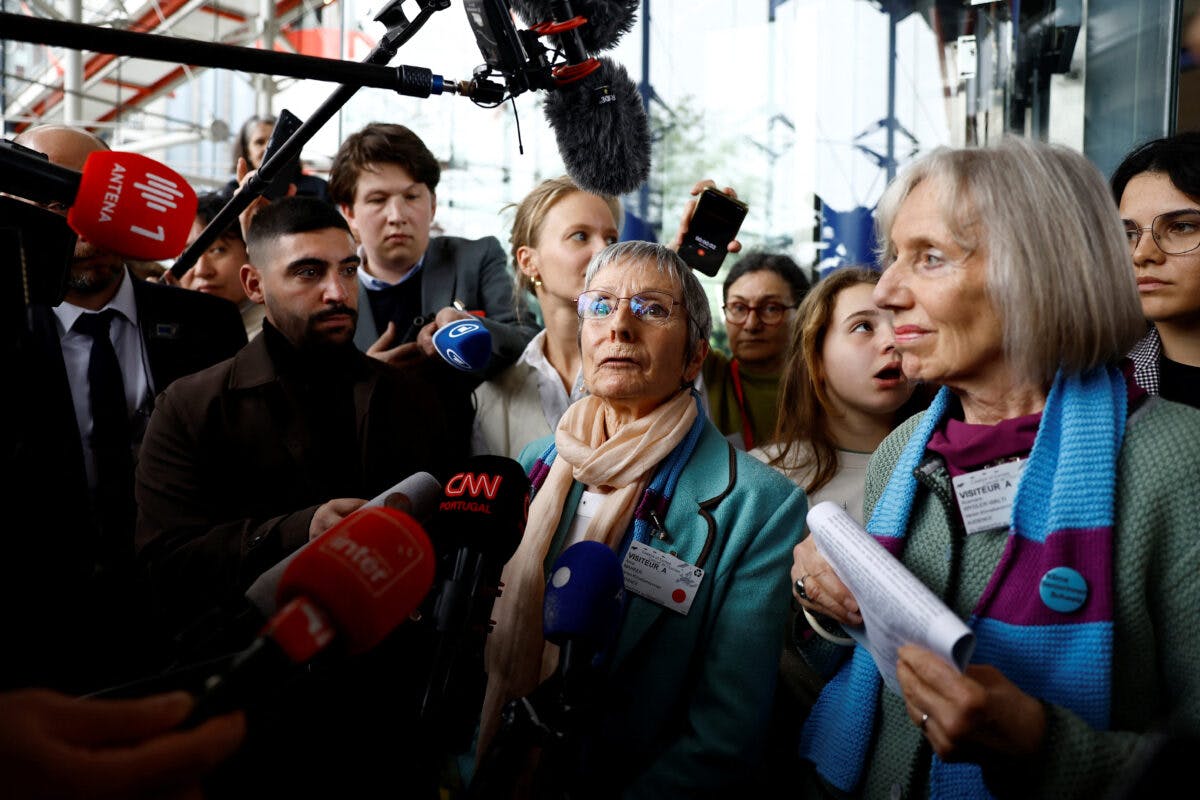 To eldre kvinner blir intervjuet av flere journalister med mikrofoner, omgitt av en folkemengde, utenfor en glassbygning.