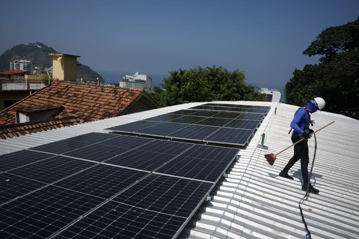 En arbeider i blå uniform og hjelm installerer solcellepaneler på et bølgepapptak med by og fjell i bakgrunnen.