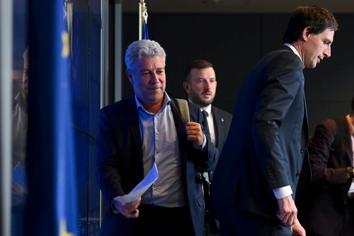 Tre menn i dress går i forskjellige retninger, hvorav en holder et papir. Et EU-flagg er delvis synlig i bakgrunnen til venstre.