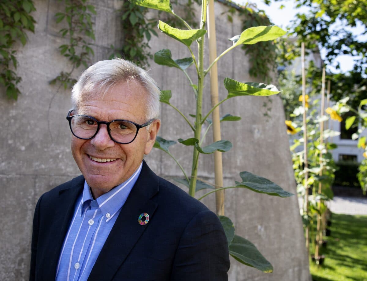 En mann med briller og dressjakke står utenfor nær en betongvegg med grønne planter som vokser i bakgrunnen.