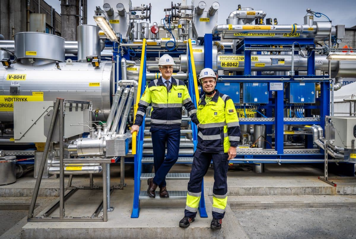 To personer som har på seg verneutstyr, inkludert hjelmer og synlige jakker, står på og i nærheten av industrimaskineri merket «Hydrogen». Den ene er i trapper, den andre på bakken, begge smilende.