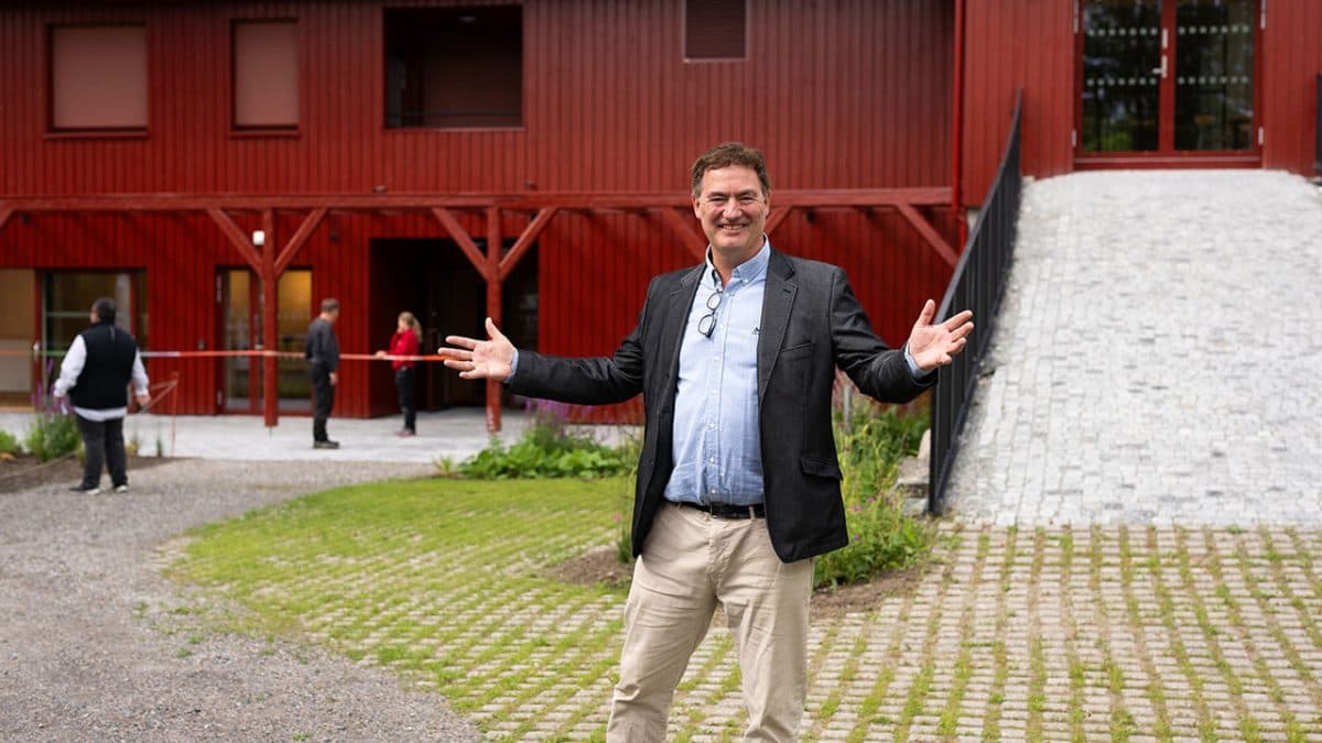 En mann i blazer og kaki står med armene utstrakt foran en rød bygning. Andre personer sees i bakgrunnen.