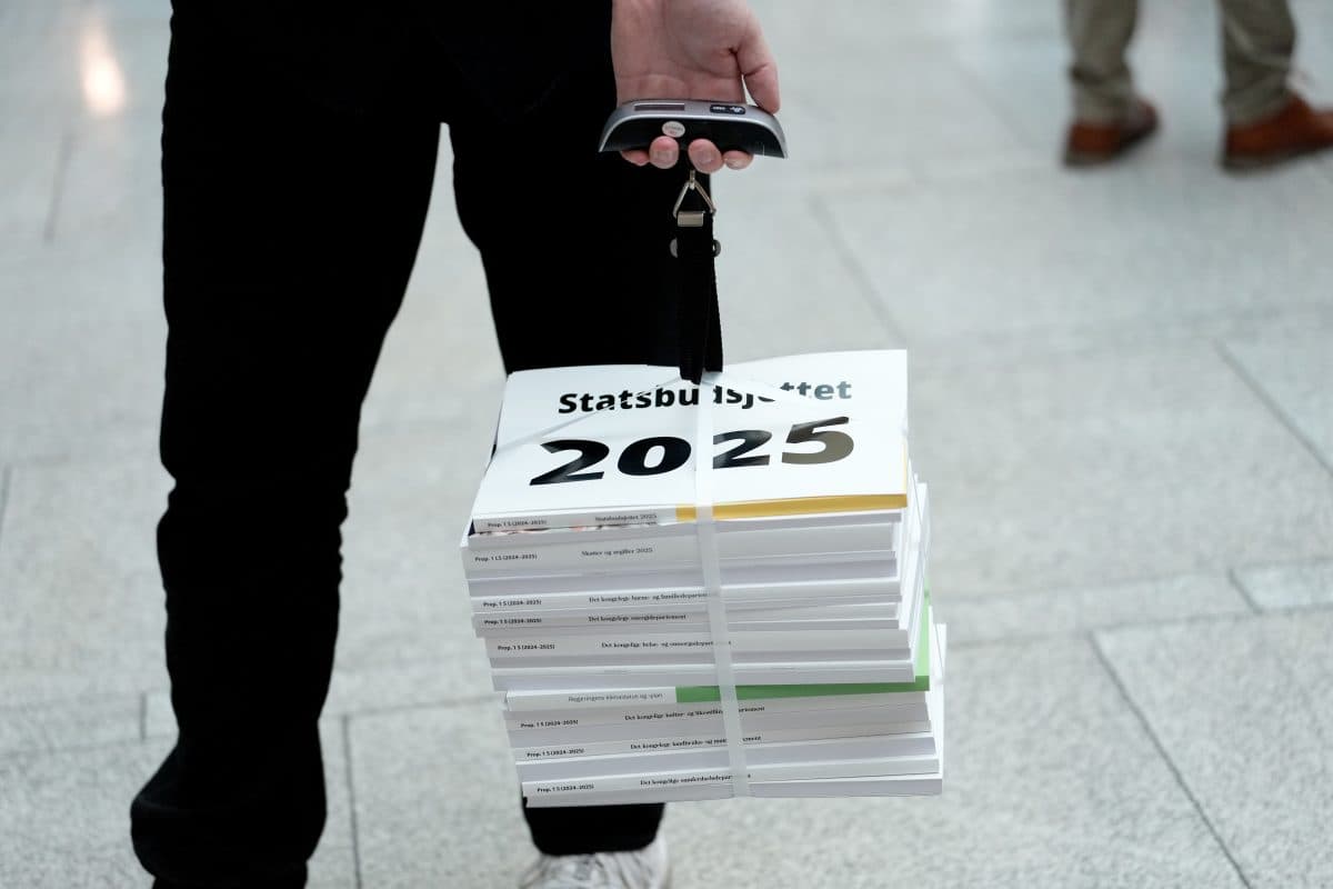 En person bærer en stor stabel med dokumenter merket «Statsbudsjettet 2025» bundet sammen, med en bagasjevekt som håndtak.