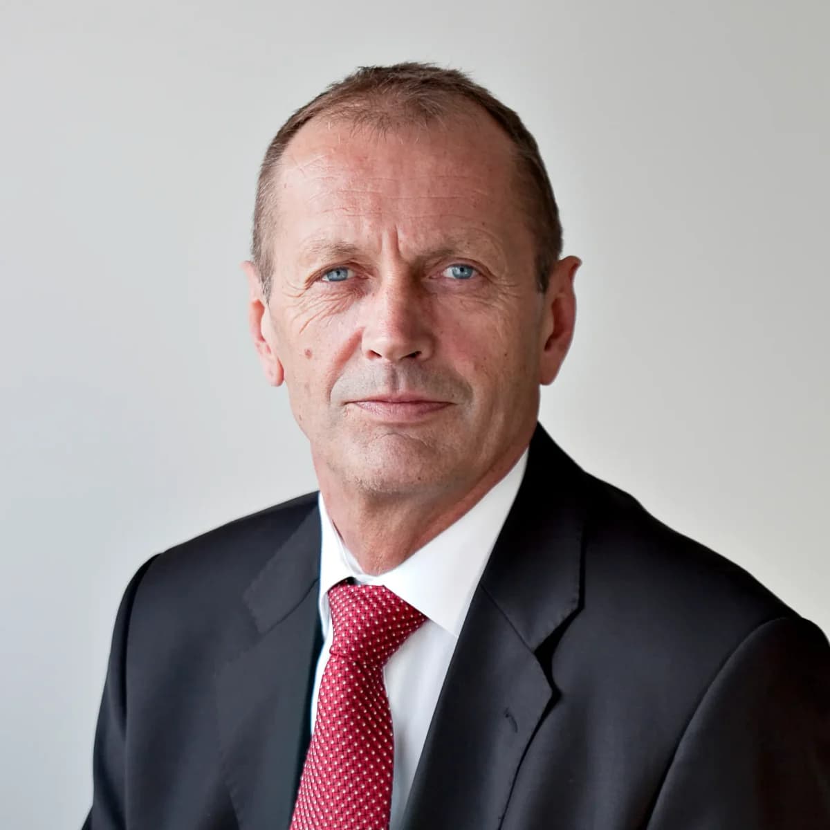 En mann i svart dress og rødt slips poserer mot en vanlig bakgrunn.