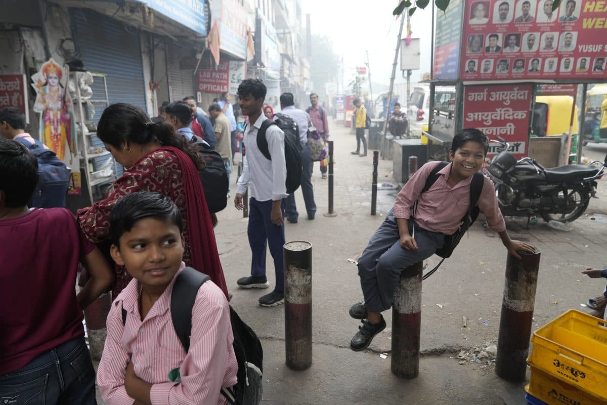 Gutter i skoleuniformer står og sitter på stolper i en travel gate i India, omgitt av andre fotgjengere og butikker.