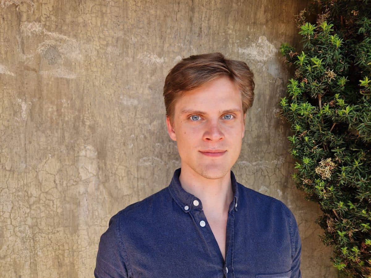 En person med kort hår i en blå skjorte står foran en strukturert vegg og grønt, og ser på kameraet.