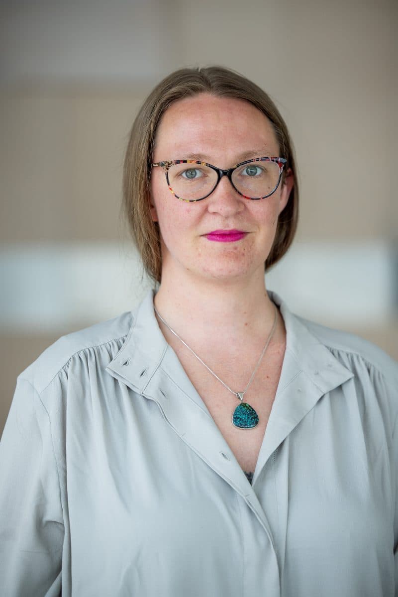 Person med briller og rosa leppestift, iført en lys grå bluse og et halskjede, stående mot en uskarp bakgrunn.