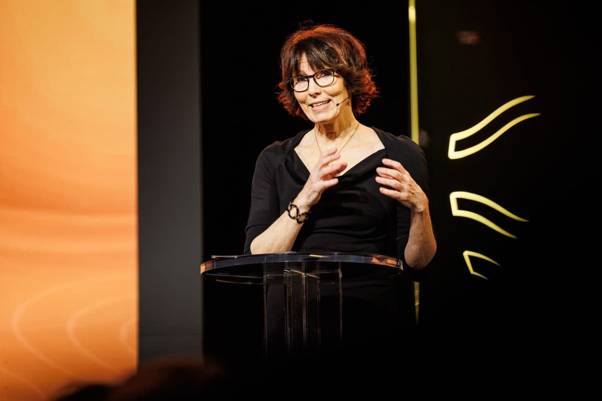 En kvinne med briller snakker ved et podium, med en oransje skjerm og bølgete gule linjer i bakgrunnen.