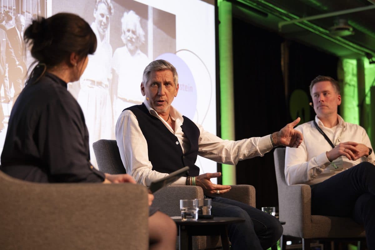 Tre personer deltok i en paneldiskusjon på scenen. En mann i midten gestikulerer mens han snakker, mens to andre lytter oppmerksomt. En projeksjon av et historisk bilde vises i bakgrunnen.