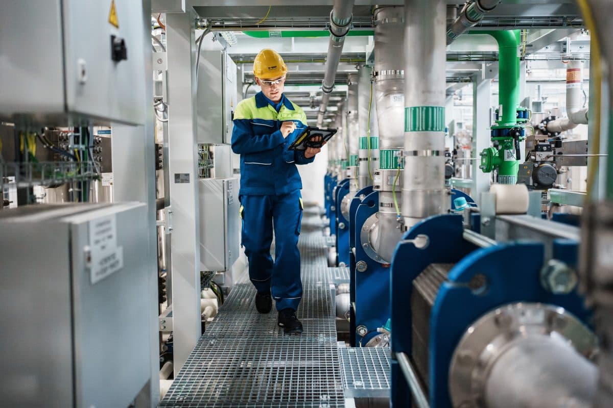En arbeider i verneutstyr inspiserer utstyr og tar notater på et nettbrett i et industrianlegg med rør og maskineri.