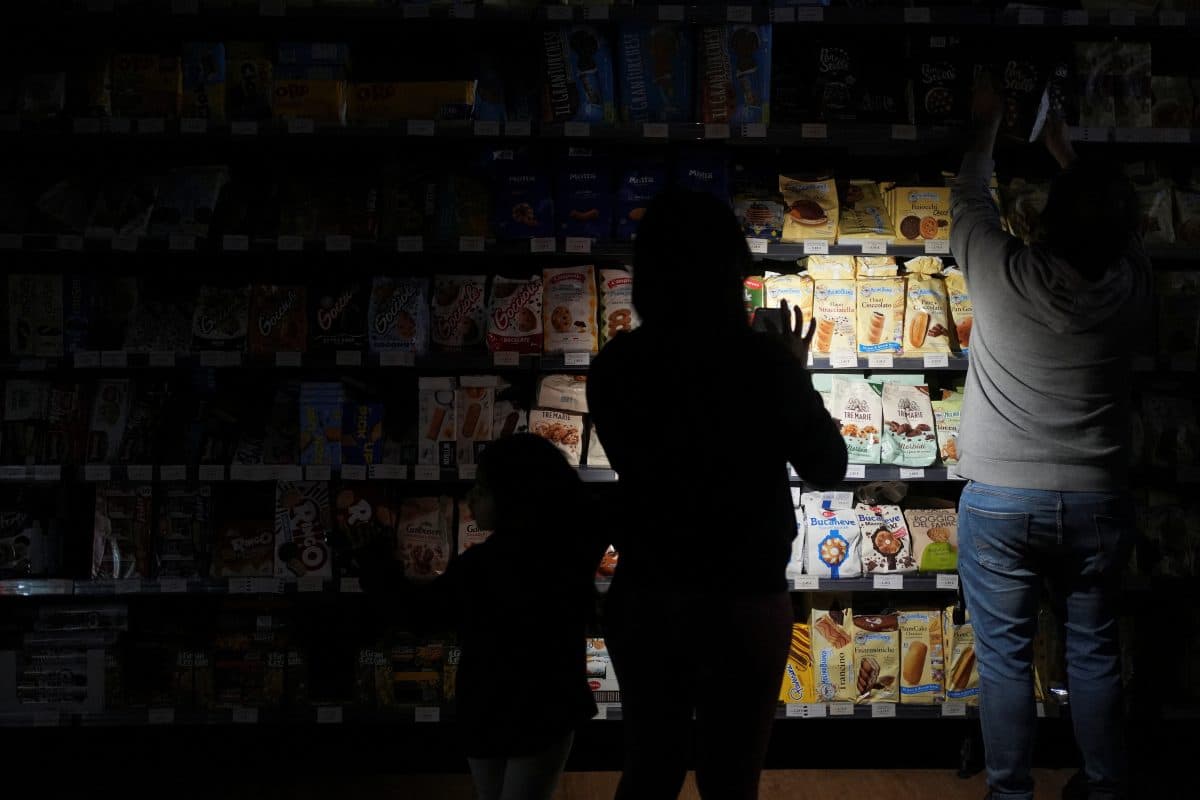 Tre personer står i en svakt opplyst midtgang i en matbutikk, med hyller med pakket mat delvis opplyst.
