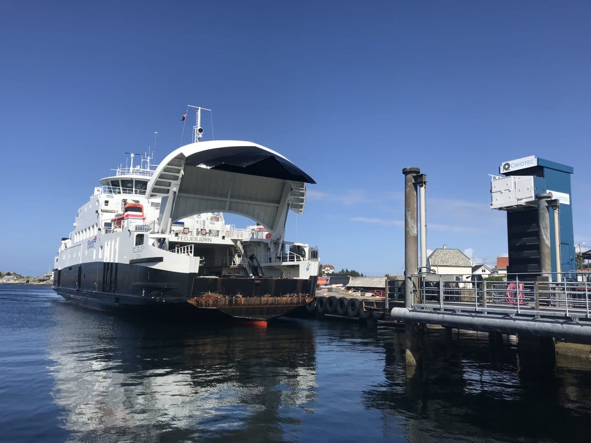 En stor hvit ferge med åpen baugrampe ligger til kai ved en brygge under en klar blå himmel, med vann og noen få bygninger synlige i bakgrunnen.
