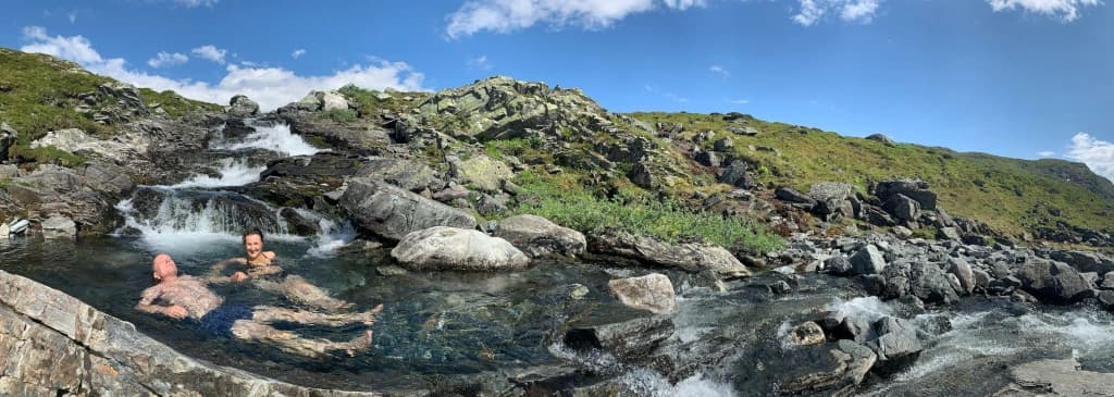 To personer slapper av i et naturlig steinbasseng i en fjellside, omgitt av rennende vann, steiner og grønne åser under en delvis overskyet blå himmel.
