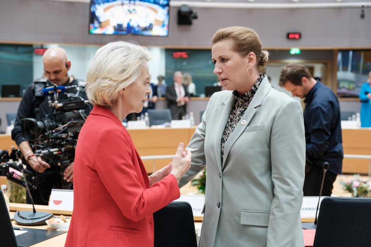 To kvinner i forretningsantrekk har en alvorlig samtale i et konferanserom, med et kamerateam og andre personer til stede i bakgrunnen.