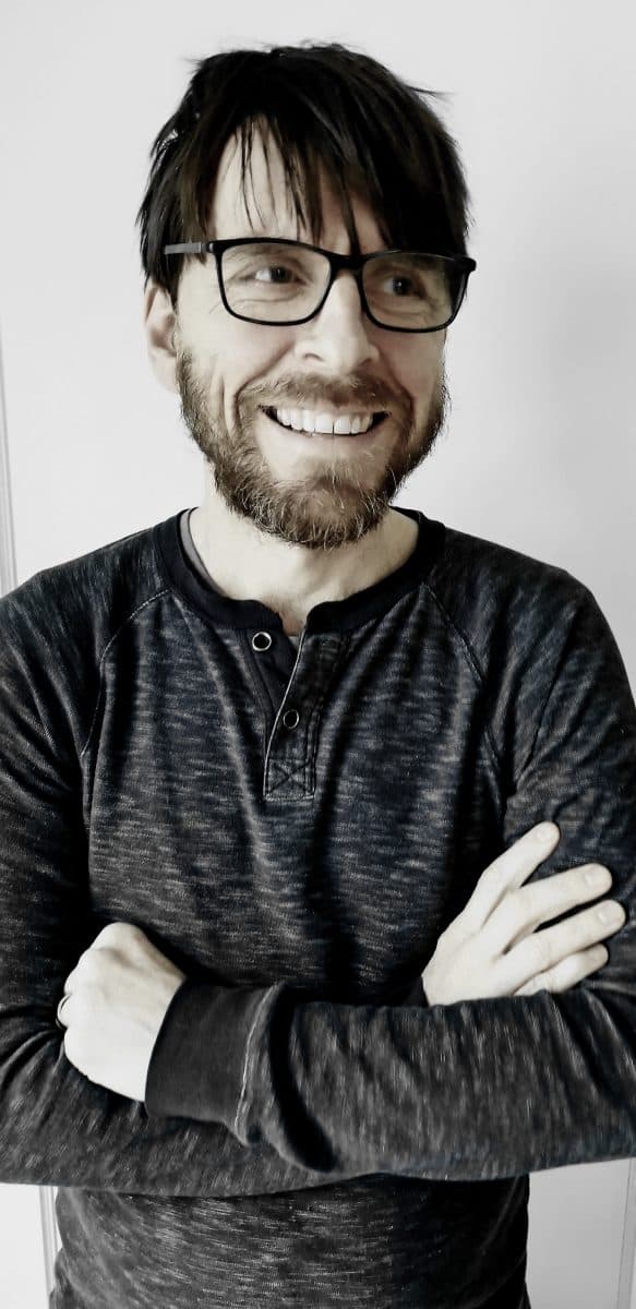 En mann med briller, skjegg og mørkt hår står smilende med armene i kors, iført en mørk langermet skjorte mot en ensfarget bakgrunn.