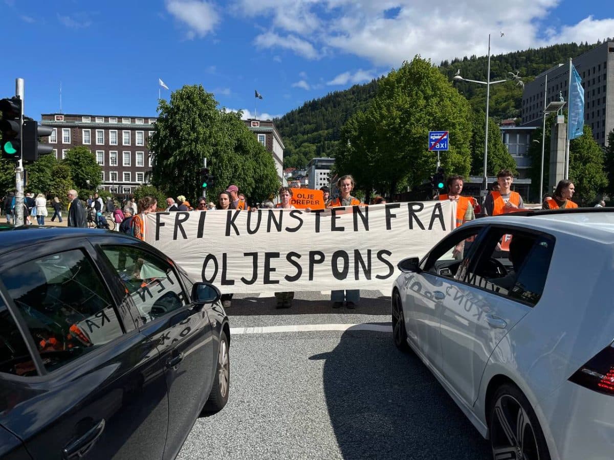 Demonstranter står i gaten og holder et stort banner med teksten «FRI KUNSTEN FRA OLJESPONS» mellom to biler, med bygninger og trær i bakgrunnen.