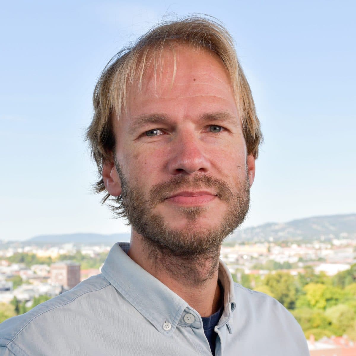 En mann med lyst hår og skjegg iført en lyseblå skjorte med knapper står utendørs med et bybilde og åser i bakgrunnen.