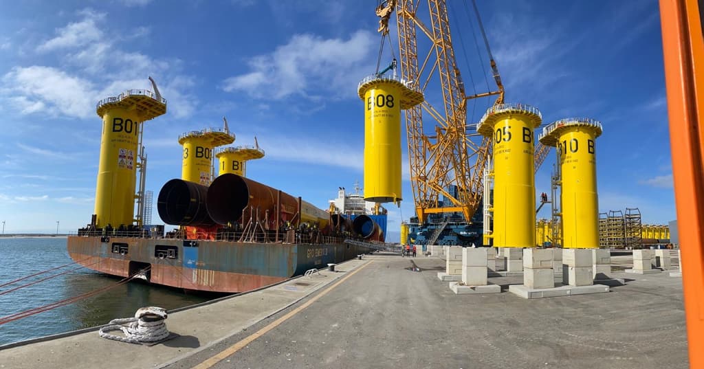 Store, gule, sylinderformede konstruksjoner merket B01 til B08 løftes med kraner fra kaikanten og over på et skip i et industrielt havneanlegg.
