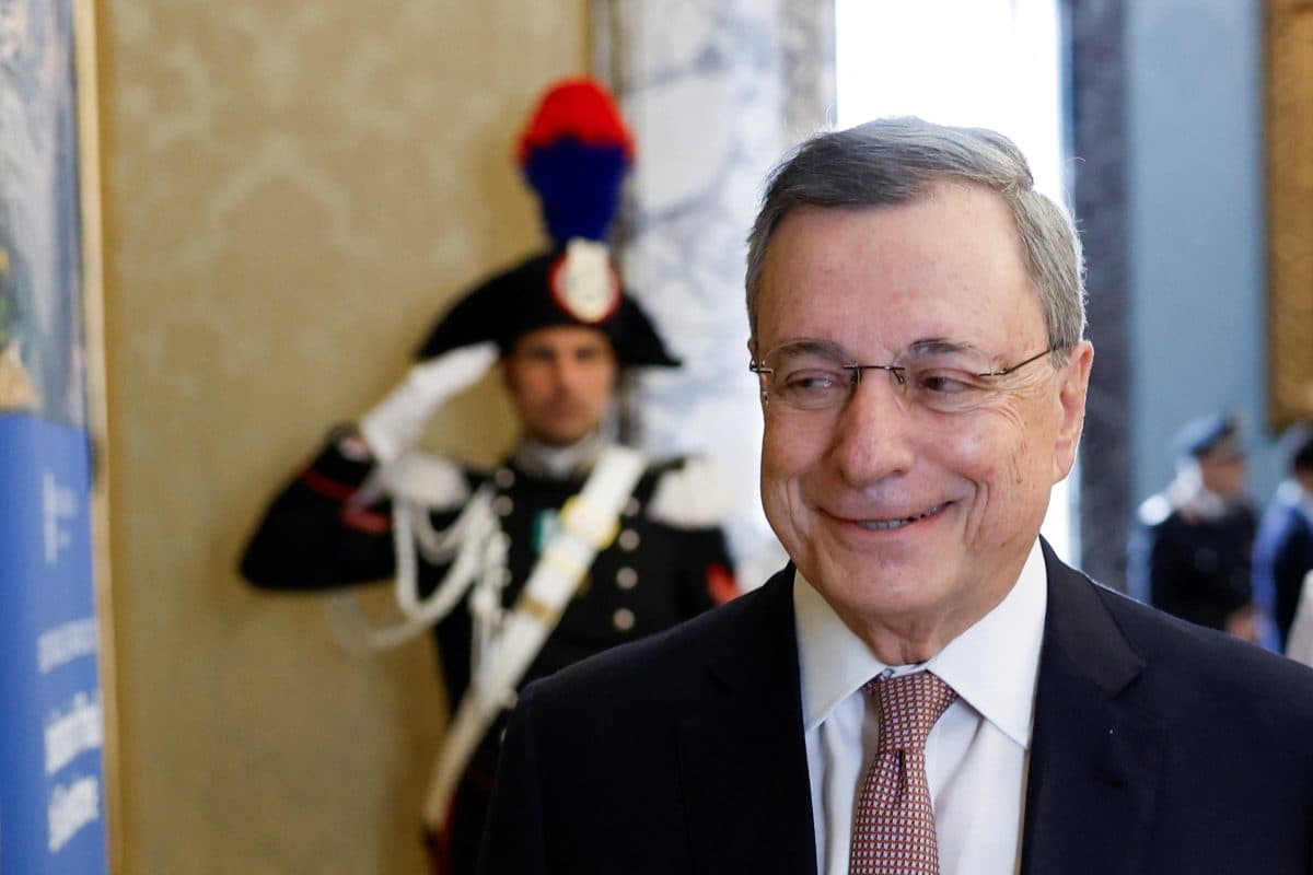 En mann i dress og slips smiler mens en uniformert vakt står i bakgrunnen og gjør honnør i et utsmykket rom.