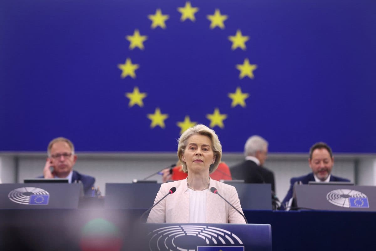 En kvinne taler på et podium med EU-flagget bak seg. Tre menn sitter ved pulter i bakgrunnen.