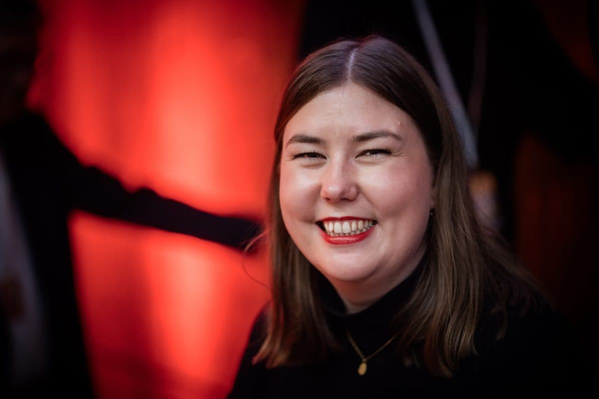 En kvinne med rett, brunt hår og svart topp smiler til kameraet, stående foran en rødbelyst bakgrunn.