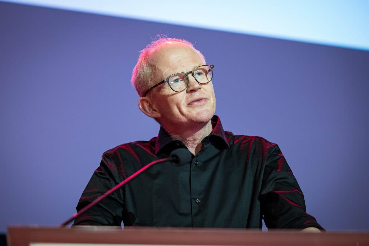 En mann med briller og svart skjorte står på et podium og snakker inn i en mikrofon mot en enkel blå og hvit bakgrunn.