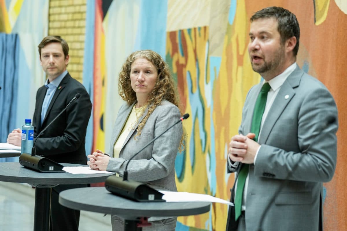 Tre personer i forretningsantrekk står ved podier med mikrofoner og snakker foran et fargerikt, abstrakt tekstilkunstverk.