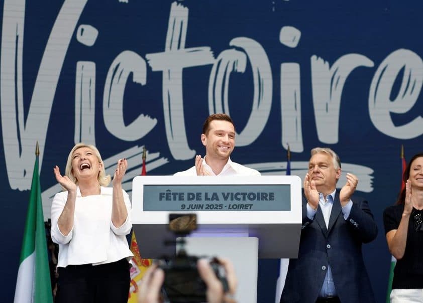 En gruppe mennesker står på scenen og smiler og applauderer foran et skilt med påskriften "Fête de la Victoire" på et arrangement.