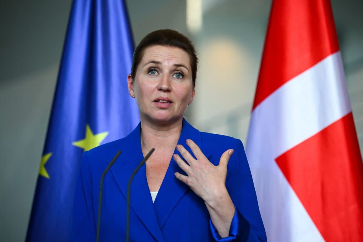 En kvinne i blå blazer taler på et podium med EU-flagg og danske flagg i bakgrunnen.