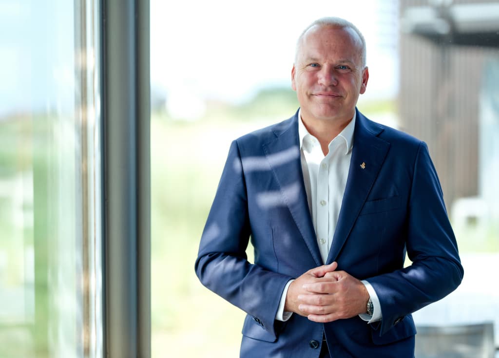 En mann i blå dress står innendørs ved et vindu og smiler med hendene foldet foran seg. I bakgrunnen skimtes sterkt lys og en utendørsscene.