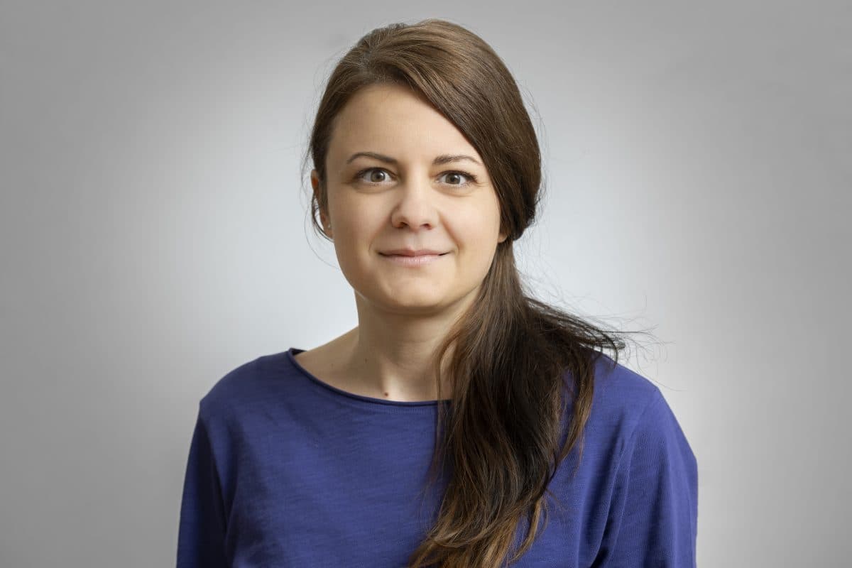 En kvinne med langt brunt hår og blå overdel står foran en vanlig, lysegrå bakgrunn og ser rett inn i kameraet.