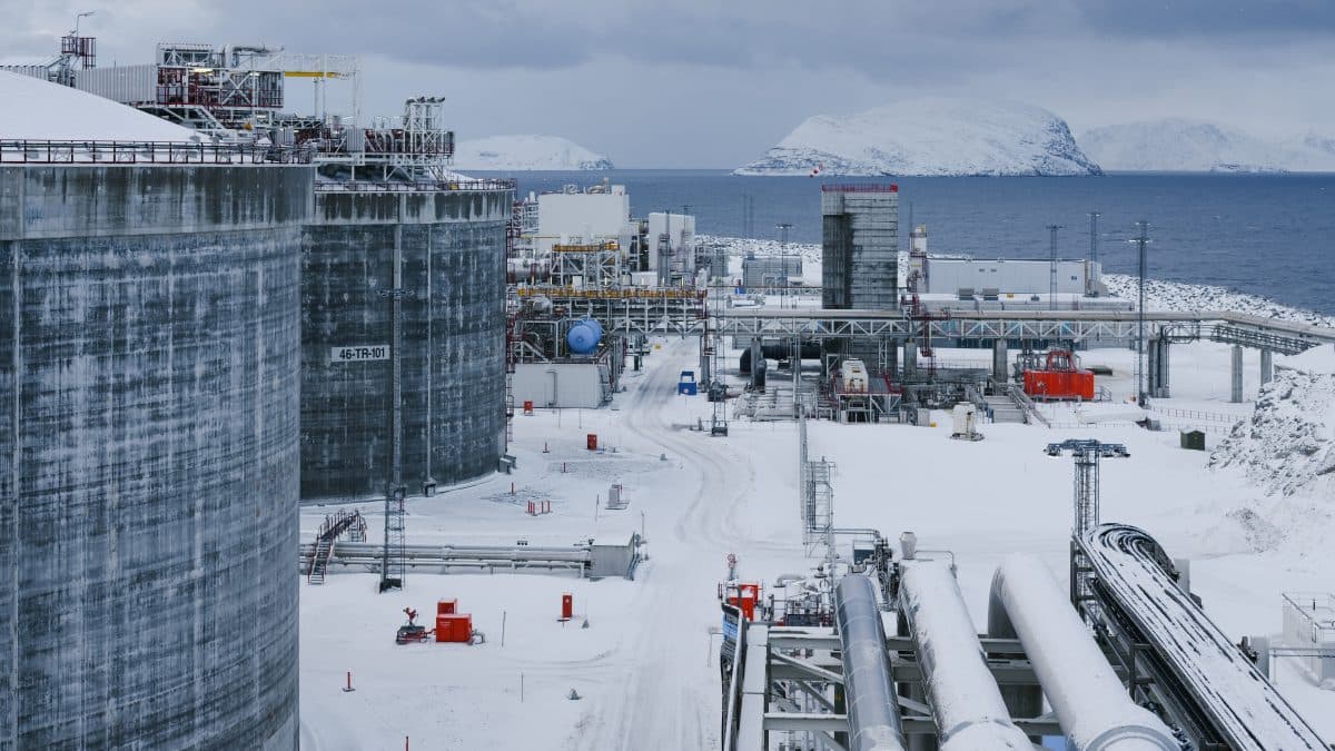 Industrianlegg med store lagertanker, rørledninger og utstyr i et snødekt landskap nær sjøen, med snødekte øyer i bakgrunnen.