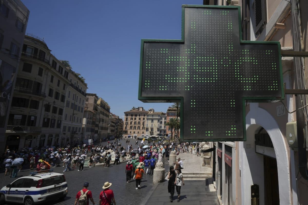 Et stort, opplyst, grønt apotek-kryssskilt med utsikt over en travel bygate med mange mennesker og historiske bygninger under en klar himmel.