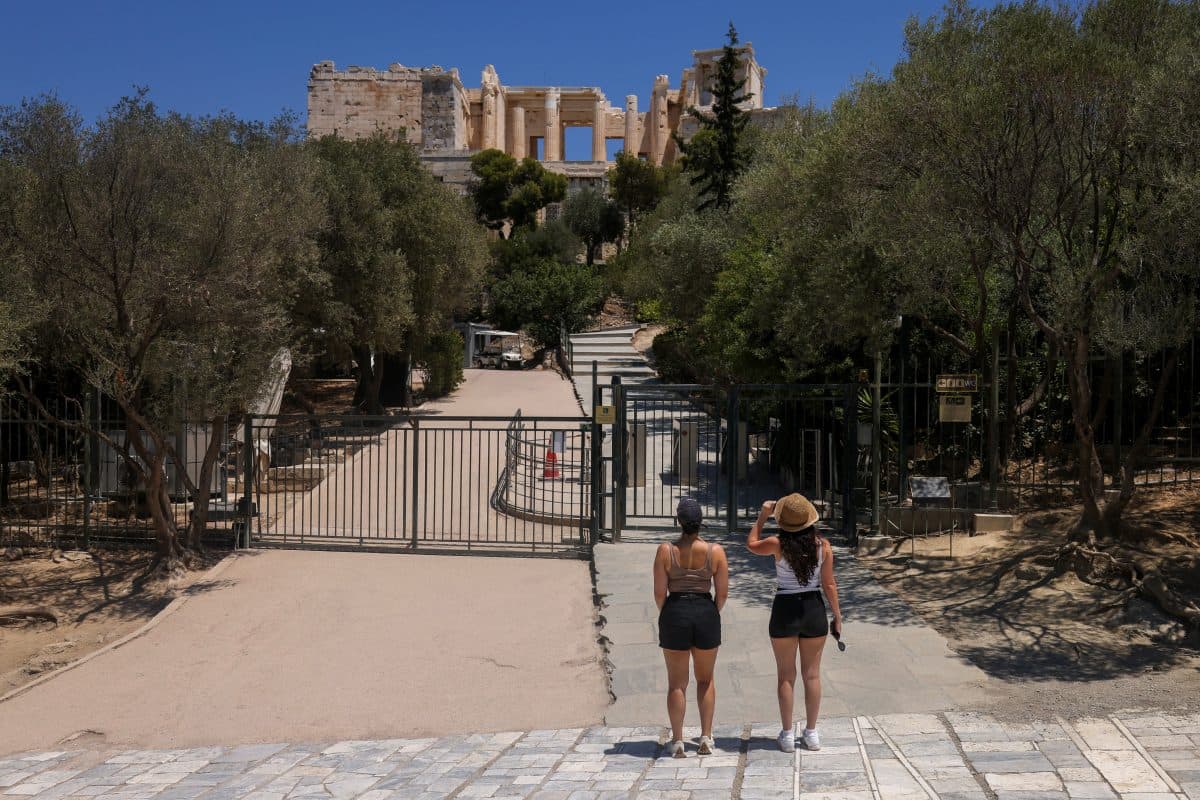 To kvinner står foran en lukket port som fører til Akropolis-ruinene i Aten, med trær langs stien og antikke bygninger i bakgrunnen.