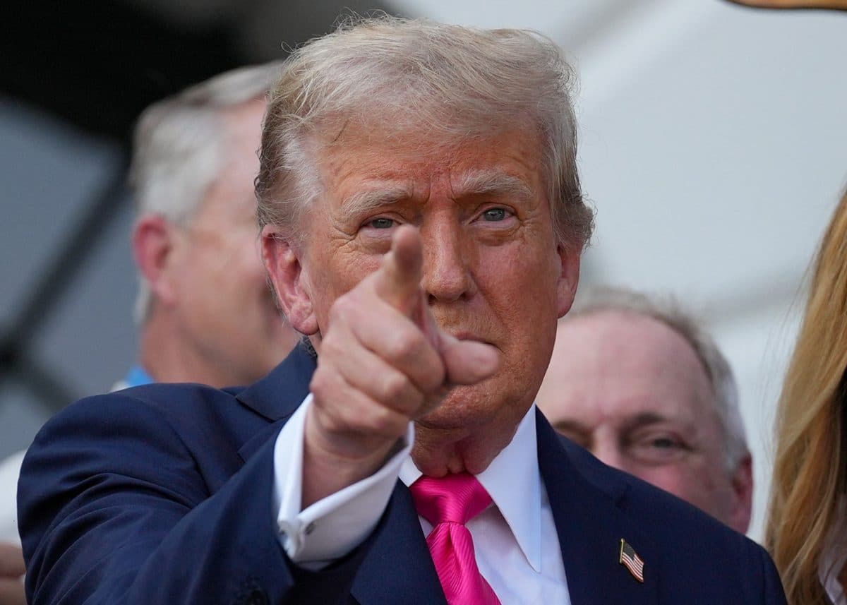 En mann i dress og rosa slips peker rett mot kameraet mens han står blant en gruppe mennesker.